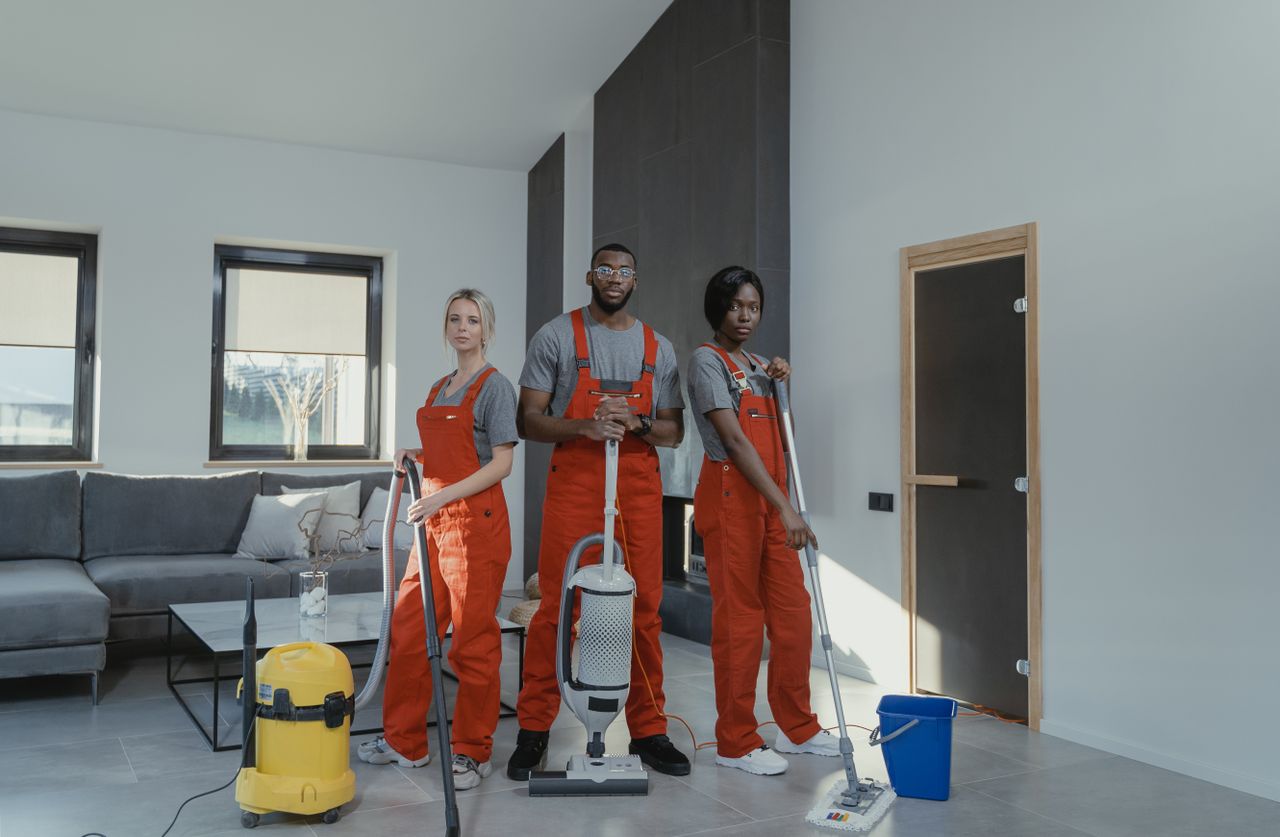 about-img-01 Diverse cleaning team in red uniforms ready to sanitize a modern living room with vacuum and mop.